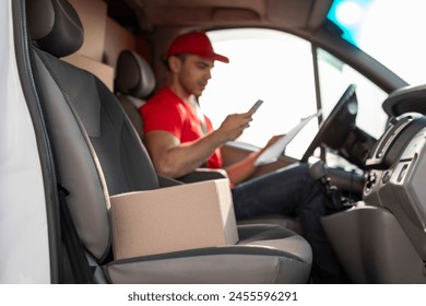 Courier man checking customer address using cellphone, sitting at drivers seat in van, focus on parcel nearby - Powered by Shutterstock