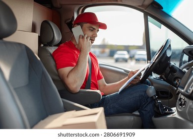 Courier man inside loaded cargo van speaking on cellphone and reviewing shipment lists and delivery route details on clipboard