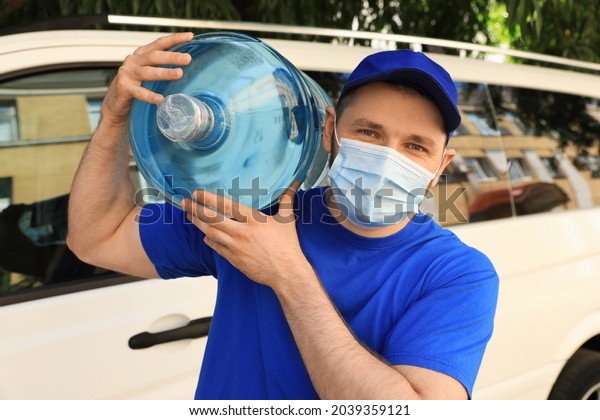 Courier in medical\
mask holding bottle of cooler water near car outdoors. Delivery\
during coronavirus\
quarantine