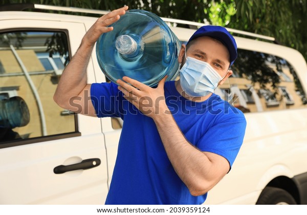 Courier in medical\
mask holding bottle of cooler water near car outdoors. Delivery\
during coronavirus\
quarantine
