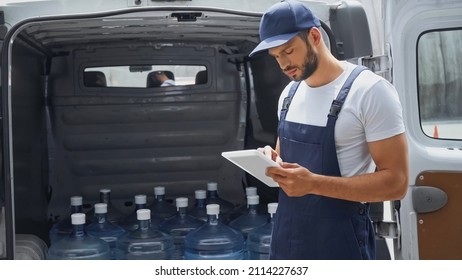 Courier in overalls using digital tablet near bottles in auto outdoors