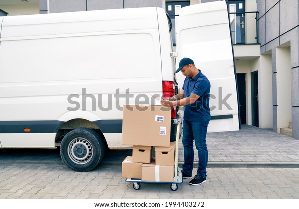 Courier with packages on a hand truck looking\
at documents