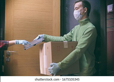 Courier in protective mask delivers parcel to client, client in medical gloves signs on tablet. Delivery service under quarantine, disease outbreak, coronavirus covid-19 pandemic conditions.