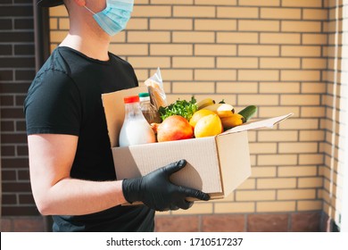 Courier In Protective Mask And Gloves Delivers Box Of Different Food. Food Delivery During Corona Virus Outbreak. Safe Home Delivery. Takeout Meal.