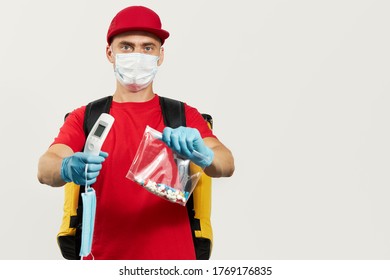 A Courier In A Red Uniform, Protective Mask And Gloves Delivers Pill Drug And Thermometer. Stay At Home And Take Care Of Your Health. Courier Drug Delivery