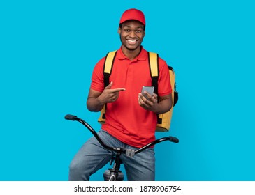 Courier Service Application. Smiling black delivery guy using smartphone and pointing at it, wearing red uniform, cap and thermo bag, riding bicycle, isolated on blue studio background
