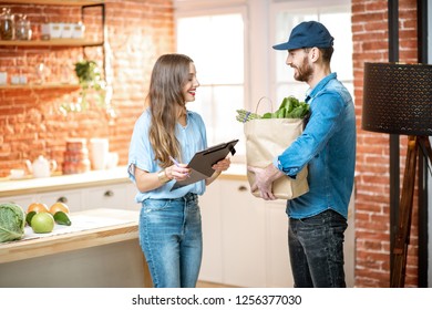 Courier Service Worker Delivering Fresh Food To A Happy Woman Client Signing Some Documents On The Kitchen At Home