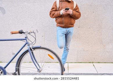 Courier using smartphone for delivery app, leaning against textured urban wall with bicycle, casual winter street style and backpack