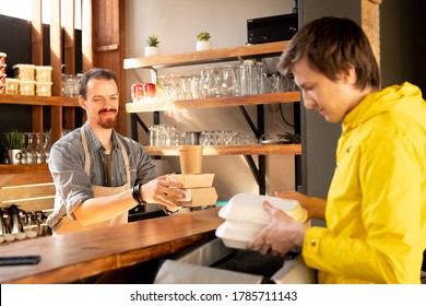 Courier In Yellow Jacket Putting Lunch Boxes Into Thermal Bag While Waiter Passing Stack Of Food Boxes To Him In Cafe