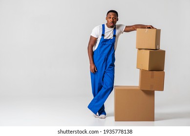 A Courier, A Young African-American Man In A Blue Jumpsuit And White T-shirt, Tired, Stands By The Paper Boxes Stacked Next To Him. Isolated On White Background. The Concept Of Delivery, Mail, Deliver