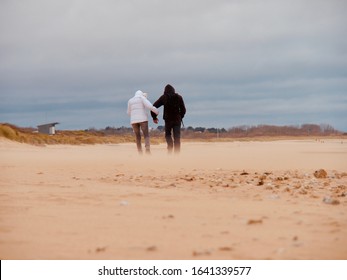 Courseulles Sur Mer, France - 09 February 2020. Couple Walking On The Beach Dressed In Jackets Windy Weather, Camera Shot From Below, Beautiful View Of Walking From Behind, Sand Flying