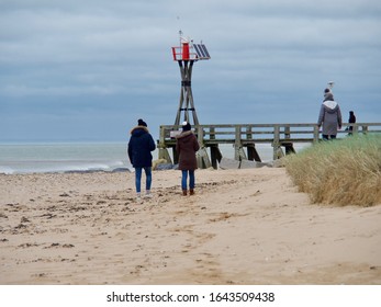 Courseulles Sur Mer, France - February 09 2020. A Couple With Children Walking On The Beach Wearing Jackets In Windy Weather, Camera Shot From Below, Flying Sand