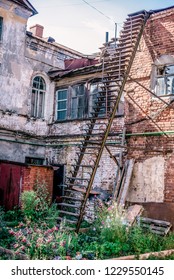 The Courtyard Of The Old Two-story Urban Brick House With  Fragment Of The Roof And Chimneys. House Of The Last Century, View Of The Windows Of The Second Floor And The Fire Escape And Flower Garden.