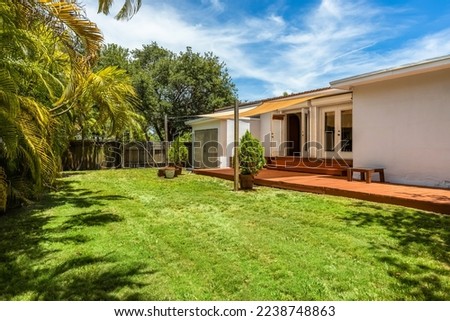 Courtyard with short grass, wooden floor, awning, wall of palm trees and wooden fence, trees and blue sky
