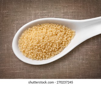 Couscous in white porcelain spoon / high-res photo of grain in white porcelain spoon on burlap sackcloth background