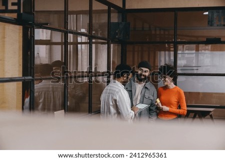 Covered peek view of young multiracial positive colleagues in casual clothes standing and looking at each other while discussing details of project in modern workspace by glass wall