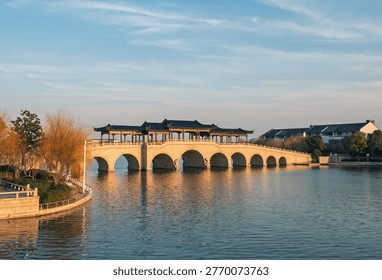 Covered stone arch bridge with multiple spans extends across a placid lake during sunset.