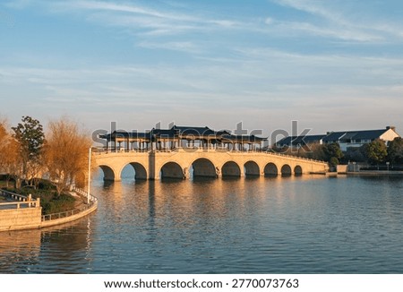 Covered stone arch bridge with multiple spans extends across a placid lake during sunset.