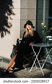 Covered Young Blond Woman In Floppy Hat And Long Sleeved Balck Dress, Sitting Behind Cafe Table Outside, Looking At The Camera.