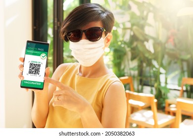 Covid 19 Digital Vaccine Passport Concept. A Beautiful And Stylish Asian Woman With Face Mask In Yellow Dress Showing The QR Code Health Pass On Her Smartphone Before Entry The Restaurant. Vaccination