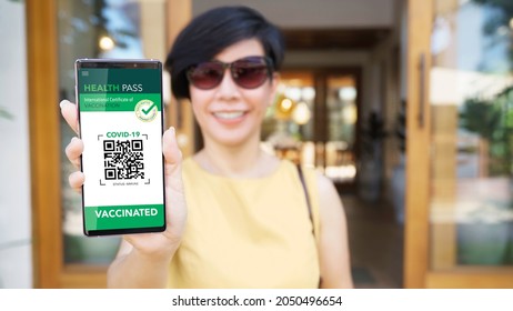 Covid 19 Digital Vaccine Passport Concept. A Beautiful And Stylish Asian Woman With Face Mask In Yellow Dress Showing The QR Code Health Pass On Her Smartphone Before Entry The Restaurant. Vaccination