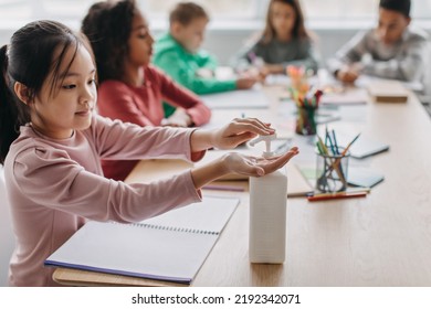 Covid 19 Prevention. Japanese Schoolgirl Disinfecting Hands At School. Kid Girl Using Sanitizer During Class Sitting With Diverse Classmates In Modern Classroom Indoors. Children Healthcare Concept