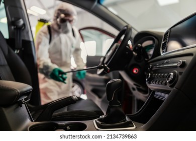 COVID 19 Situation Warning. A Man In A Protective White Uniform And A Sprayer Prevents The Spread Of The Infection Inside Of A Car, Showroom. Stay Responsible, Wear The Mask, Coronavirus, Virus