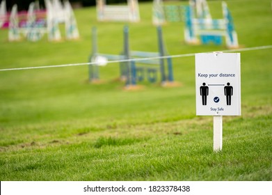 A Covid 19 Social Distancing Sign Staked Into Grass With An Horse Jumps At An Equestrian Show Jumping Outdoor Event In The Background.