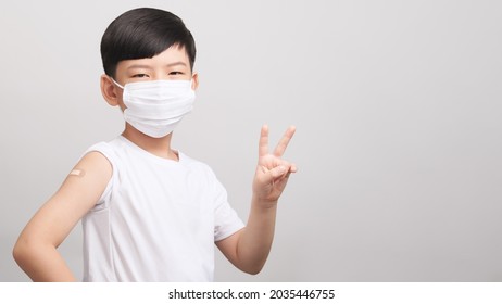 COVID 19 Vaccines For Kids Concept. Studio Portrait Of An Adorable Asian Boy With Medical Face Mask Do V Sign Hand Gesture After Got His First Dose Vaccine. Immune, Safe, Available, Back To School.