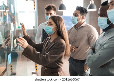 Covid Outbreak And Workplace. Side View Of Diverse Multiethnic Group Of Business People In Protective Masks Having Meeting In Board Room, Brainstorming, Using Glass Wall With Sticky Post-it Notes