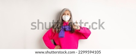 Covid, pandemic and social distancing concept. Cheerful asian senior woman in respirator showing thumbs-up, wearing face mask from coronavirus, white background.