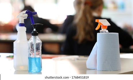 Covid Safe School. Inside A Classroom With Cleaning Products On Display Such As Hand Sanitizer, Disinfectant, Paper Towel On A Desk. Corona Virus Issues And Impact. Deliberate Blurred Students