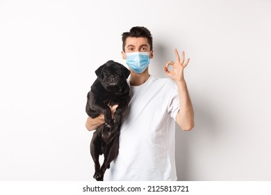 Covid-19, animals and quarantine concept. Young man in medical mask holding cute black pug dog, showing okay sign, like and approve, standing over white background - Powered by Shutterstock