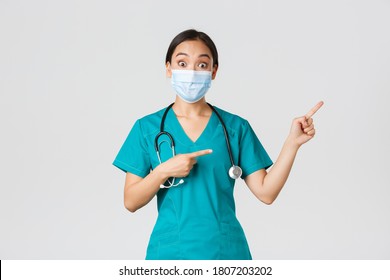 Covid-19, Coronavirus Disease, Healthcare Workers Concept. Enthusiastic Happy Asian Female Doctor, Physician Or Nurse In Medical Mask And Scrubs, Pointing Upper Right Corner, Show Awesome Promo