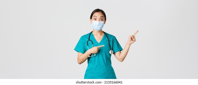 Covid-19, Coronavirus Disease, Healthcare Workers Concept. Enthusiastic Happy Asian Female Doctor, Physician Or Nurse In Medical Mask And Scrubs, Pointing Upper Right Corner, Show Awesome Promo