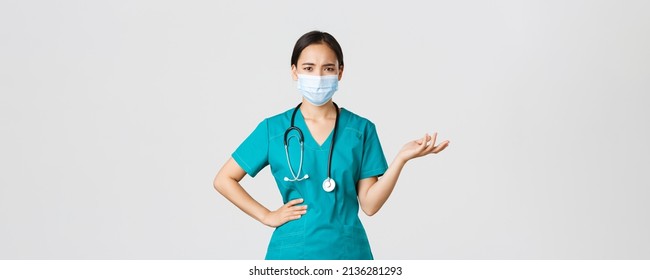 Covid-19, coronavirus disease, healthcare workers concept. Confused and upset asian female doctor in medical mask and scrubs, raise hand up puzzled, cant understand what problem, white background - Powered by Shutterstock