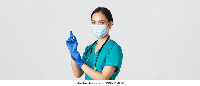 Covid-19, coronavirus disease, healthcare workers concept. Professional confident asian female doctor, nurse in medical mask and scrubs, put on rubber gloves for examination, white background - Powered by Shutterstock