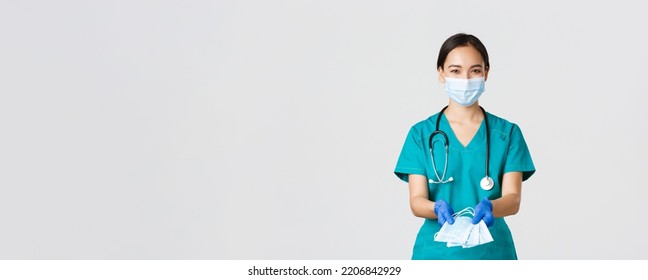 Covid-19, Coronavirus Disease, Healthcare Workers Concept. Friendly Smiling Asian Physician, Female Doctor In Scrubs And Rubber Gloves Handing Over Medical Masks To Patients, White Background