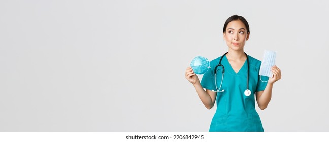 Covid-19, Coronavirus Disease, Healthcare Workers Concept. Thoughtful Smiling Asian Female Doctor, Physician In Scrubs Looking Dreamy Upper Left Corner, Holding Respirator And Medical Mask