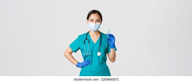 Covid-19, Coronavirus Disease, Healthcare Workers Concept. Smiling Confident Asian Female Nurse In Scrubs And Medical Mask, Holding Syringe With Vaccine, Doing Vaccination Shot, White Background