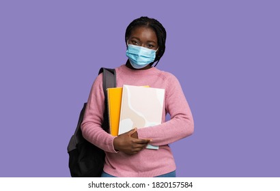 Covid-19 And Education Concept. Black Female University Student Wearing Medical Mask, Holding Workbooks And Backpack, Looking At Camera While Posing Over Purple Studio Background, Copy Space