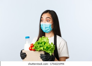 Covid-19, Grocery Store, Employment, Small Business And Preventing Virus Concept. Surprised Asian Female Shop Worker, Cashier In Medical Mask And Gloves Raise Eyebrows Amused, Hold Bag With Groceries