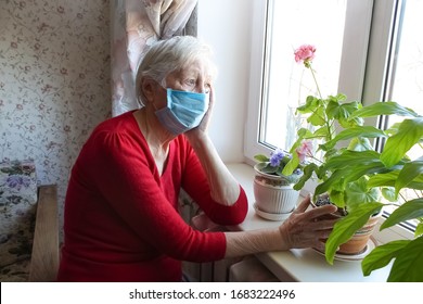 The Covid-19, health, safety and pandemic concept - senior old lonely woman wearing protective medical mask sitting near the window at home for protection from virus
