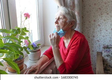 The Covid-19, Health, Safety And Pandemic Concept - Senior Old Lonely Woman Wearing Protective Medical Mask Sitting Near The Window At Home For Protection From Virus