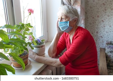 The Covid-19, Health, Safety And Pandemic Concept - Senior Old Lonely Woman Wearing Protective Medical Mask Sitting Near The Window At Home For Protection From Virus