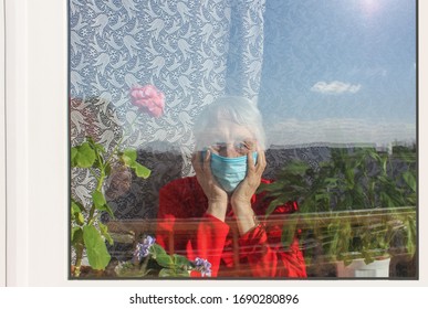 The Covid-19, Health, Safety And Pandemic Concept - Senior Old Lonely Woman Wearing Protective Medical Mask Sitting Near The Window At Home For Protection From Virus