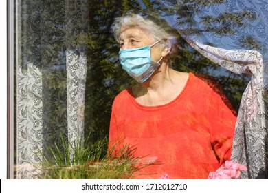 The Covid-19, Health, Safety And Pandemic Concept - Senior Old Lonely Woman Wearing Protective Medical Mask Sitting Near The Window At Home For Protection From Virus