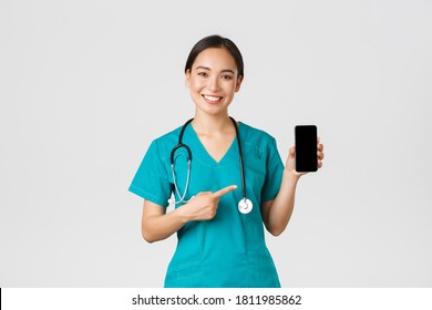 Covid-19, healthcare workers and online medicine concept. Smiling asian female doctor, physician in scrubs pointing finger at mobile screen, recommend download checkup, consultation application