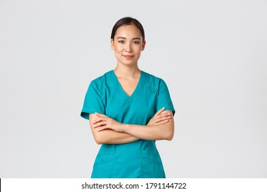 Covid-19, healthcare workers, pandemic concept. Confident smiling asian nurse in scrubs standing self-assured, cross arms chest. Professional doctor ready for surgery, standing white background