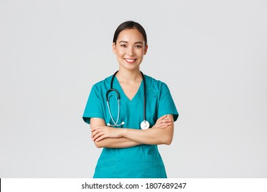 Covid-19, healthcare workers, pandemic concept. Portrait of confident smiling, attractive asian female nurse in scrubs, with stethoscope, cross arms chest and looking at camera, white background